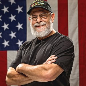 Bearded Vietnam veteran standing in front of an American Flag with arms crossed